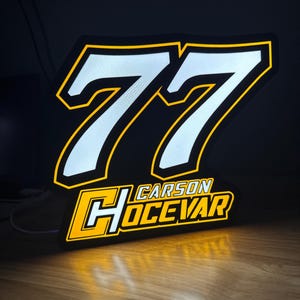 May include: Illuminated sign featuring the number 77 in white with a yellow outline, set against a black background. The name "CARSON HOCEVAR" is displayed below in a yellow and white design. The sign is lit with a warm glow.