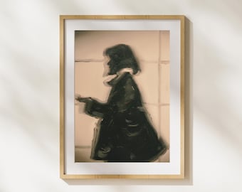 May include: A framed art print featuring a blurred, impressionistic painting of a person in a dark coat. The artwork has a muted color palette with a beige background and a light wooden frame.