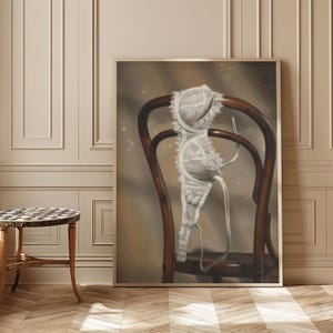 以下が含まれることがあります： 暗い木製の椅子の背もたれに掛けられた白いレースのブラジャーが描かれた額入りのアートワーク。ブラジャーには複雑なレースのディテールがあります。背景は、落ち着いたニュートラルなカラーパレットです。