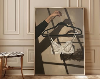 May include: A framed painting depicts a white lace bra hanging on a black hanger. The bra is centered, with delicate lace detailing. The background is a neutral beige, with shadows suggesting sunlight. The painting is displayed in a room with a small table.