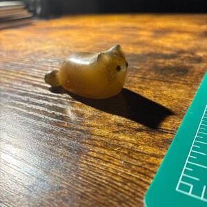 Puede incluir: Pequeña figura de gato de color ámbar con ojos negros y cuerpo redondeado. El gato está tumbado, con la cola enrollada hacia un lado. La figura está sobre una superficie de madera.
