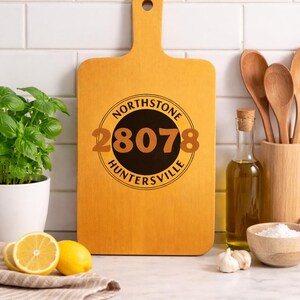 May include: A wooden cutting board with a handle, featuring a circular design with the text "NORTHSTONE 28078 HUNTERSVILLE". The board is surrounded by kitchen items, including lemons, basil, and olive oil.