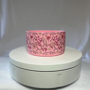 May include: Pink cylindrical container with a repeating pattern of pink flamingos, flowers, and the text "GET YOUR PINK BACK". The container is sitting on a white rotating platform. The background is a light gray.