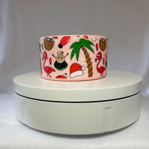Pode incluir: Um recipiente cilíndrico rosa com um padrão repetitivo de ilustrações festivas. O design inclui um boneco de neve, palmeiras, flamingos, chapéus de Papai Noel e cocos. O recipiente está em uma plataforma giratória branca.