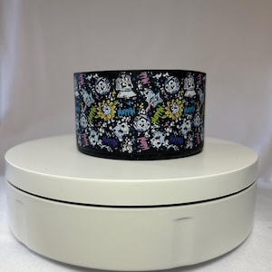 May include: A black cylindrical container with a colorful cartoon pattern. The design includes smiling suns, skulls, and other characters. The word "Muffin" is also visible in the design. The container is sitting on a white rotating display.