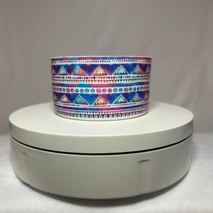 May include: A cylindrical container with a colorful geometric pattern in shades of blue, purple, pink, and orange. The design features triangles, dots, and lines. The container sits on a white rotating platform.