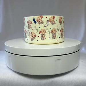 May include: A cylindrical, off-white ceramic mug featuring a repeating pattern of a cartoon character, socks, and stars. The mug is displayed on a white, rotating platform. The design includes a variety of colors, including red, blue, and green.