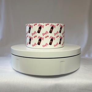 May include: A cylindrical container with a repeating pattern of Dr. Pepper bottles, pink bows, and cherries on a white background. The container is sitting on a white rotating platform.