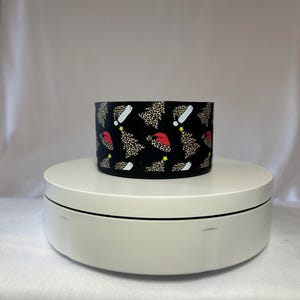 Op de afbeelding: Een zwart cilindrisch object met een herhaald patroon van kerstbomen, kerstmutsen en witte uitroeptekens. De kerstbomen en kerstmutsen hebben een luipaardprint. Het object staat op een wit draaiend platform.