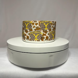Op de afbeelding: Een cilindrische keramische container met een herhalend patroon van luipaardprint, bruine koeienvlekken en gele "Y"-vormen. De container is wit en staat op een witte, roterende displaystandaard.