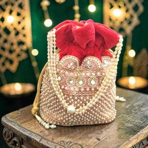 May include: A drawstring pouch bag featuring a bright red velvet top section and a light pink base adorned with intricate gold embroidery, small inset mirrors, and a dense pattern of white faux pearls, complete with a long pearl-beaded handle.