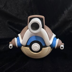 May include: A spherical figurine resembling a Blastoise-themed Poké Ball, featuring a tan lower half, a blue and white upper section with a central white button, and a prominent gray cannon protruding from the top, flanked by two smaller side cannons.