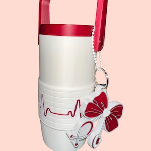 May include: White and red insulated tumbler with a red handle and a decorative stethoscope charm. The tumbler features a red heartbeat line design. The charm is a red bow with a stethoscope design.