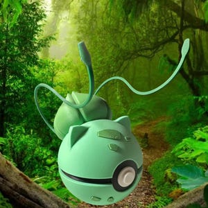 May include: A mint green, spherical device with a black and white circular accent, and two curved, light green tendrils extending upwards. The device has a cartoon-like design with small, rounded features and is set against a lush, green forest background.