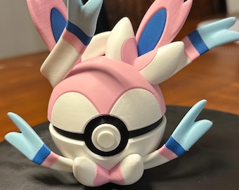 May include: A 3D-printed figure of a Sylveon Pokémon. The figure features a pink, white, and blue color scheme. The figure has multiple ribbon-like appendages and a spherical body. The figure is sitting on a black surface.