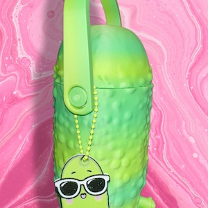 May include: A novelty green pickle-shaped container with a matching handle and a small charm of a pickle wearing sunglasses. The container has a textured surface and a small green stem. The background is pink and white.