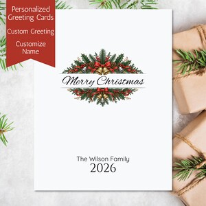 May include: A white greeting card featuring a central illustration of green pine branches, red holly berries, pinecones, and gold bells tied with red ribbons. The text Merry Christmas is printed in black, followed by The Wilson Family and 2026.