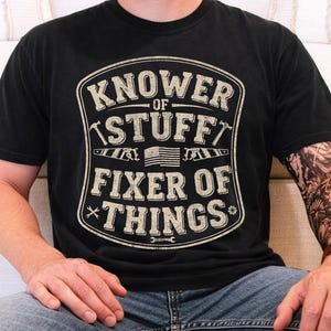 May include: Black t-shirt with a distressed graphic that reads "Knower of Stuff, Fixer of Things." The design includes tools and an American flag. The shirt is worn by a person wearing blue jeans.