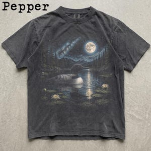 May include: A charcoal grey short-sleeve t-shirt featuring the word Pepper in the top left corner. The centre graphic depicts a loon swimming on a lake at night under a full moon, with a starry sky, pine trees, and white water lilies on the water.