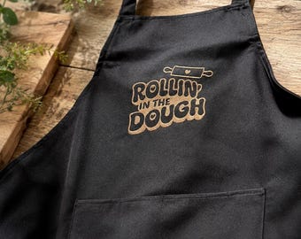 May include: A black apron featuring a neck strap and a front pocket. The chest area displays an embroidered design in a metallic gold color, consisting of a rolling pin icon with a small heart and the text "ROLLIN' IN THE DOUGH" in a bold font.
