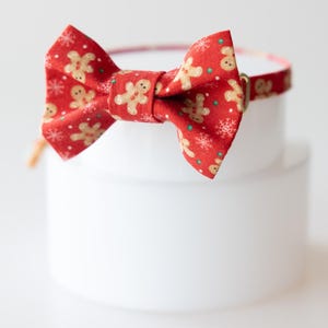 May include: A red fabric bow tie featuring a repeating pattern of light brown gingerbread men and white snowflakes. The bow tie is attached to a matching thin fabric collar strap with a small metallic gold-toned buckle adjustment piece.