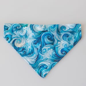 May include: A triangular pet bandana featuring a swirling ocean wave pattern in shades of blue and white. The fabric is printed with a watercolor-style design, creating a dynamic and flowing visual effect. The bandana is designed to be worn around a pet's neck.