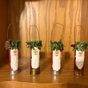 May include: Four handmade Christmas ornaments shaped like Santa Claus. Each ornament is made from a red and brass shell casing, with a painted Santa face and white beard. They are decorated with greenery and a wire handle.