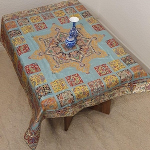 May include: A rectangular, light blue tablecloth with a central starburst design and a repeating pattern of colourful squares. Two small blue and white vases sit on the tablecloth. The tablecloth is draped over a small wooden table.