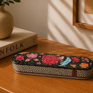 Könnte beinhalten: Eine längliche, ovale Schachtel mit einem schwarzen Deckel, der ein lebhaftes, mehrfarbiges Blumenmuster mit rosa, roten und orangefarbenen Blüten sowie einem blauen Vogel zeigt. Die Seiten der Schachtel sind mit einem geometrischen Sternmuster verziert.