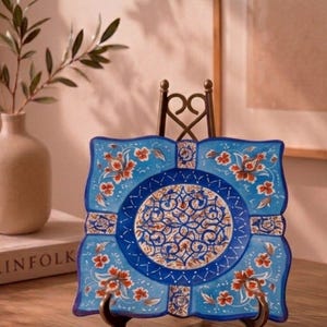 May include: A square ceramic plate featuring a vibrant blue background with intricate white and orange floral patterns. The center displays a detailed circular medallion with swirling motifs. The plate is held upright by a dark metal display stand.