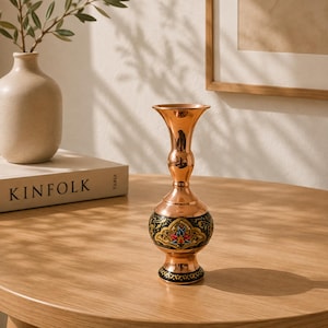 Può includere: Un vaso in rame lucido con collo sottile e bordo svasato. La base arrotondata presenta una fascia nera decorata con intricati motivi floreali nei colori rosso, blu e oro. Il vaso poggia su una base circolare con finiture nere.