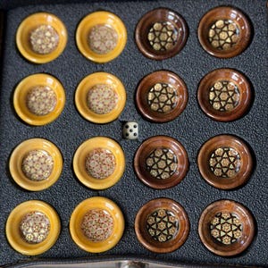 May include: A set of sixteen circular game pieces arranged in a grid within a black foam tray. Eight pieces are light yellow wood with intricate geometric inlays, and eight are dark brown wood with gold-toned star patterns. A single die is included.