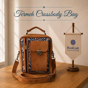 Könnte beinhalten: Eine braune Umhängetasche aus Wildleder mit Tragegriff und einem blauen Stoffeinsatz mit Muster. Die Tasche hat eine vertikale Reißverschlusstasche, eine Frontklappe mit Metallverschluss und einen verstellbaren Schultergurt. Text: Termeh Crossbody Bag.
