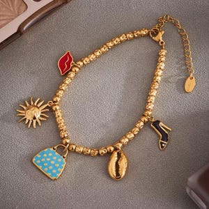 Peut inclure: Bracelet à breloques doré avec divers charms décoratifs. Les breloques comprennent un soleil, un sac à main à pois bleus, un coquillage, un escarpin et des lèvres rouges. Le bracelet est posé sur une surface grise.