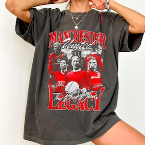 May include: Dark grey t-shirt with a graphic of Manchester United players in red. The text "MANCHESTER United" and "The Red Devils LEGACY" are in red. The shirt has a vintage, worn appearance.