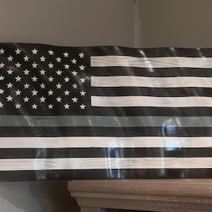 May include: A wooden American flag with a distressed finish. The flag features white stars on a black field, black and white stripes, and a gray stripe. The flag is mounted on a wooden surface and has a wavy design.