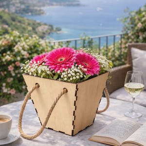 Może przedstawiać: Drewniany kosz na kwiaty z uchwytami z liny, wypełniony żywymi różowymi gerberami i białą gipsówką. Kosz stoi na marmurowym stole z kawą, winem i otwartą książką, z widokiem na morze.