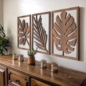 Op de afbeelding: Drie houten wandpanelen met botanische ontwerpen. Elk paneel heeft een andere bladvorm uitgesneden uit donker hout, tegen een witte achtergrond en omlijst in bijpassend hout. De panelen worden boven een houten kast weergegeven.