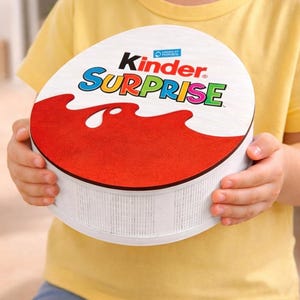 Peut inclure: Une boîte ronde Kinder Surprise tenue par une personne. Le couvercle blanc porte l'inscription "Kinder Surprise" en couleurs et un motif de vague rouge. La boîte est blanche avec des lignes verticales.