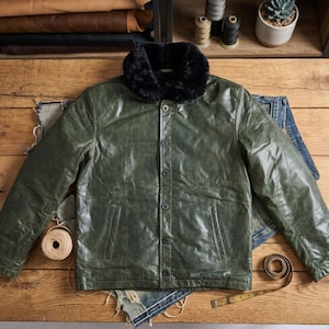 May include: A green leather jacket with a black fur collar and button closure. The jacket has two front pockets and is displayed on a wooden surface with denim fabric and sewing supplies.