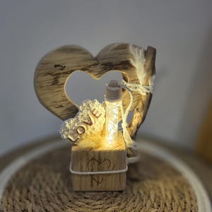 Peut inclure: Décoration en forme de cœur en bois avec une petite bouteille lumineuse au centre. Le mot "LOVE" est visible sur un élément décoratif. La base est un petit bloc de bois avec un cœur sculpté. L'ornement est sur une surface tissée.