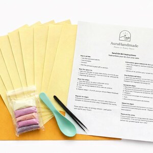 Puede incluir: Kit de arte de arena Aura Handmade con instrucciones. El kit incluye hojas de papel amarillo, una bolsa de arena de colores, una cuchara turquesa y una herramienta negra. Las instrucciones est&aacute;n impresas en una hoja blanca con el logotipo de Aura Handmade.