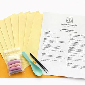 May include: Sand art kit with instructions. Includes yellow paper sheets, a bag of colored sand, a teal spoon, and a black tool. The instructions are printed on a white sheet with the Aura Handmade logo.