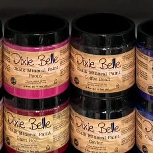 May include: A shelf displays multiple jars of Dixie Belle Chalk Mineral Paint. The jars have black lids and labels with the brand name and product information. The paint colors vary, including shades of pink, blue, and neutral tones.