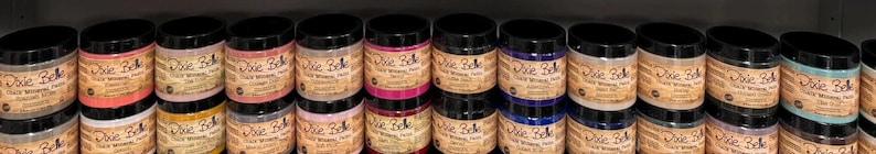 May include: A shelf displays multiple jars of Dixie Belle Chalk Mineral Paint. The jars have black lids and labels with the brand name and product information. The paint colors vary, including shades of pink, blue, and neutral tones.