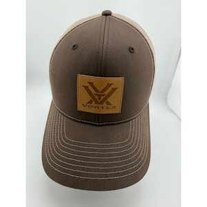 May include: A brown baseball cap with a mesh back and a tan square patch on the front. The patch features the Vortex logo and the text VORTEX in dark brown. The cap has a curved brim with light brown stitching and a button on the top crown.