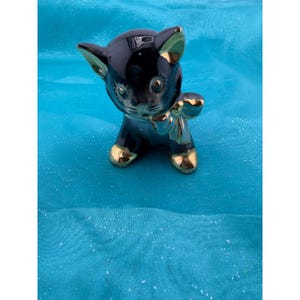 May include: A glossy black ceramic cat figurine featuring gold-painted accents on the ears, paws, and a bow around the neck. The cat has a smooth, reflective surface with painted facial features including eyes, a nose, and whiskers in a seated pose.