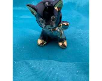 May include: A glossy black ceramic cat figurine featuring gold-painted accents on the ears, paws, and a bow around the neck. The cat has a smooth, reflective surface with painted facial features including eyes, a nose, and whiskers in a seated pose.