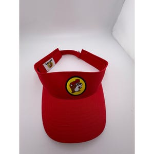 May include: A red visor with a curved brim and a strap. The visor features a circular patch with a cartoon beaver in a yellow circle. The text "Buc-ee's" is printed on the strap.