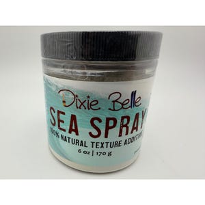 May include: A clear plastic jar with a black lid, labeled "Dixie Belle Sea Spray." The label features a wave design and text indicating it's a 100% natural texture additive. The jar contains 6 oz / 170 g of product.